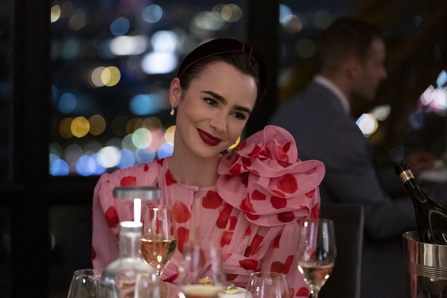 Emily in Paris Season 3: Lily Collins as Emily Cooper sitting at a dinner table surrounded by wine glasses and smiling as she wears a pink and red ruffled dress.