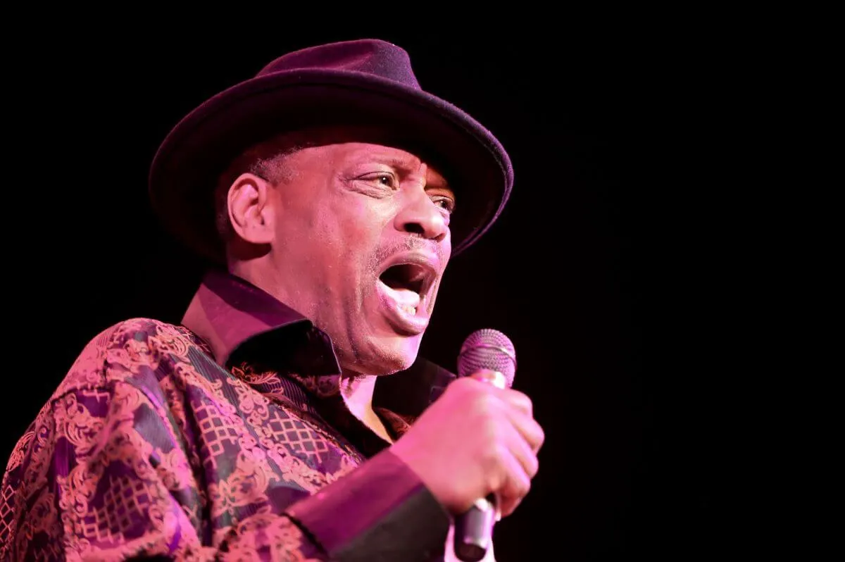 Alexander O'Neal, who just finished up a farewell tour, wears a patterned shirt and a fedora. He sings into a microphone.