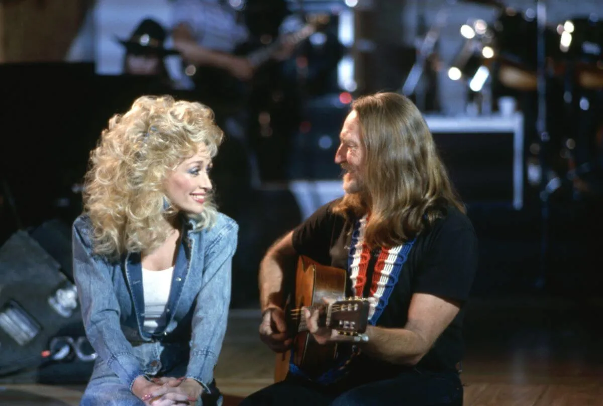 Dolly Parton and Willie Nelson sit next to each other on a stage. He plays a guitar.