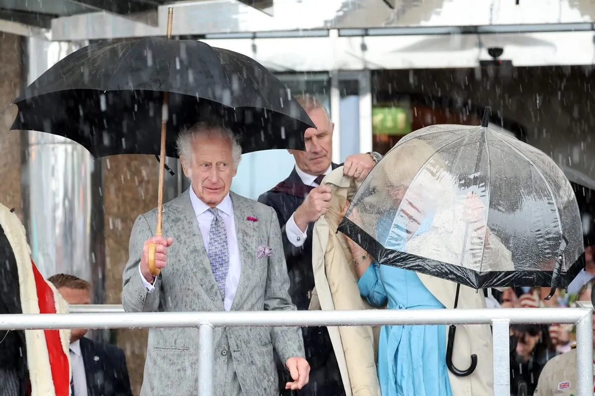 King Charles and Queen Camilla holding umbrella while an aide assist Camilla during visit to St. Helier, Jersey